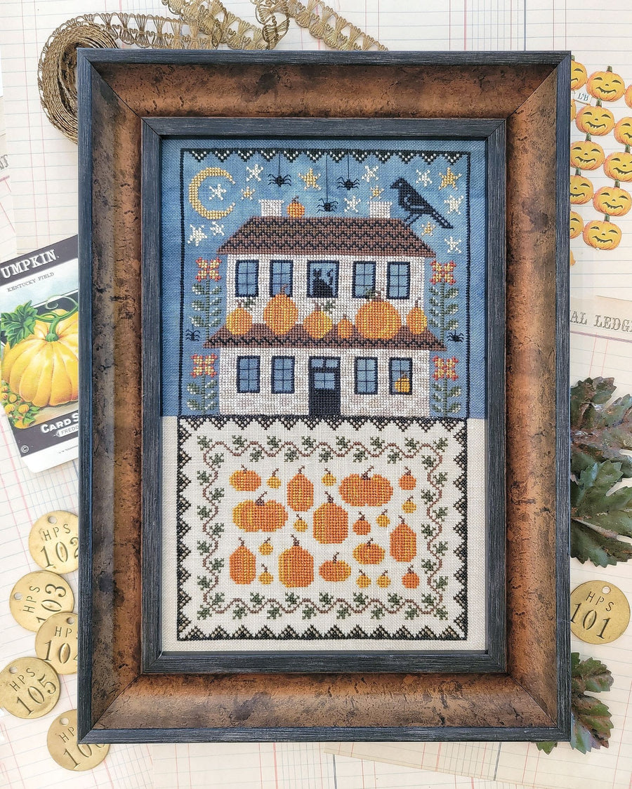 Framed cross-stitched piece with a house and pumpkins on a blue background.