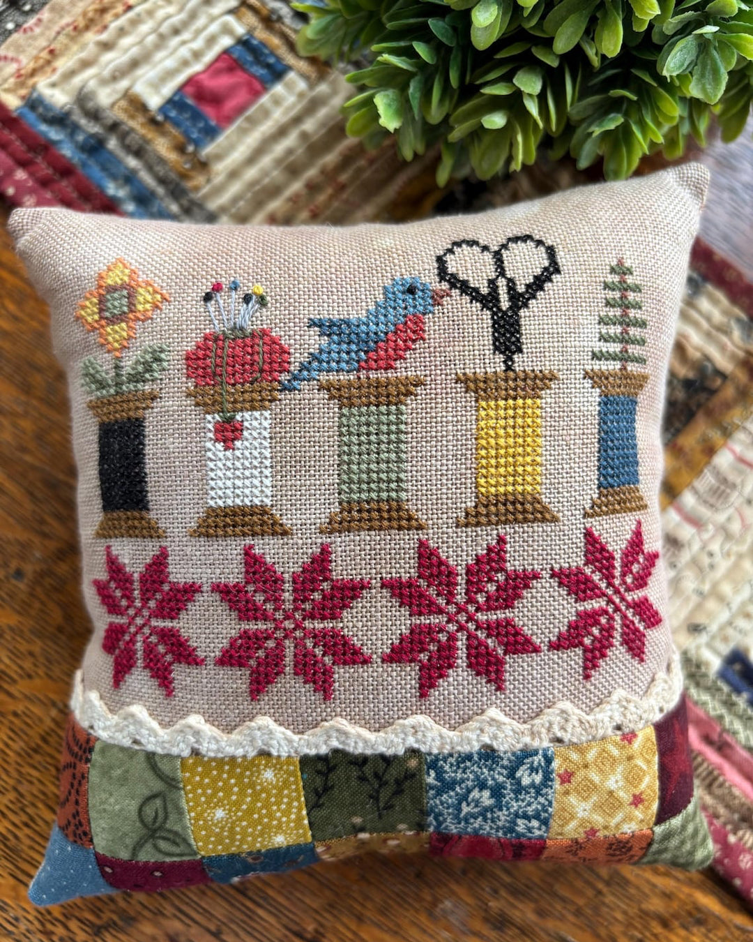 A square decorative throw pillow with a beige linen front featuring a colorful cross-stitch pattern of flowers, birds, scissors, and trees, with a red snowflake border and a patchwork fabric hem, resting on a wooden surface.
