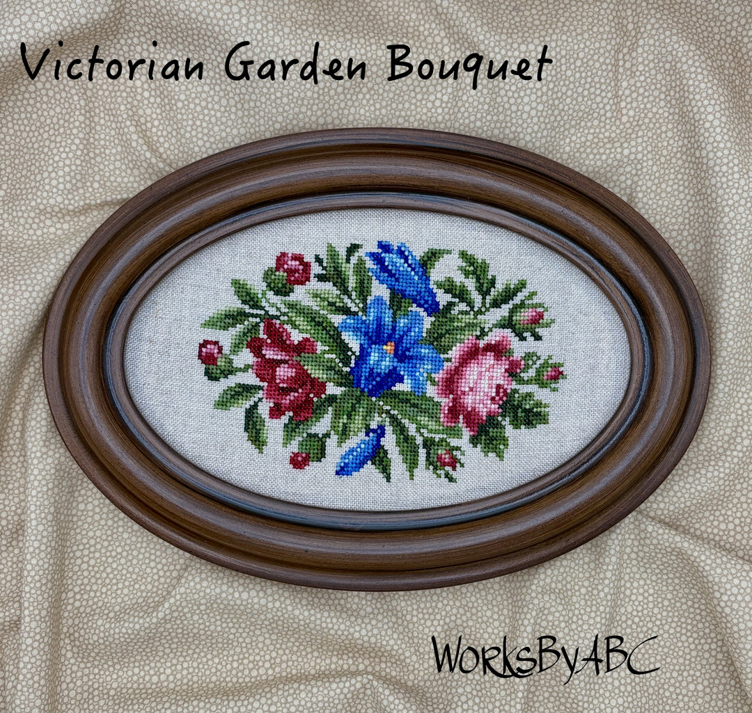 Framed cross-stitched floral design labeled 'Victorian Garden Bouquet' on a textured fabric background.