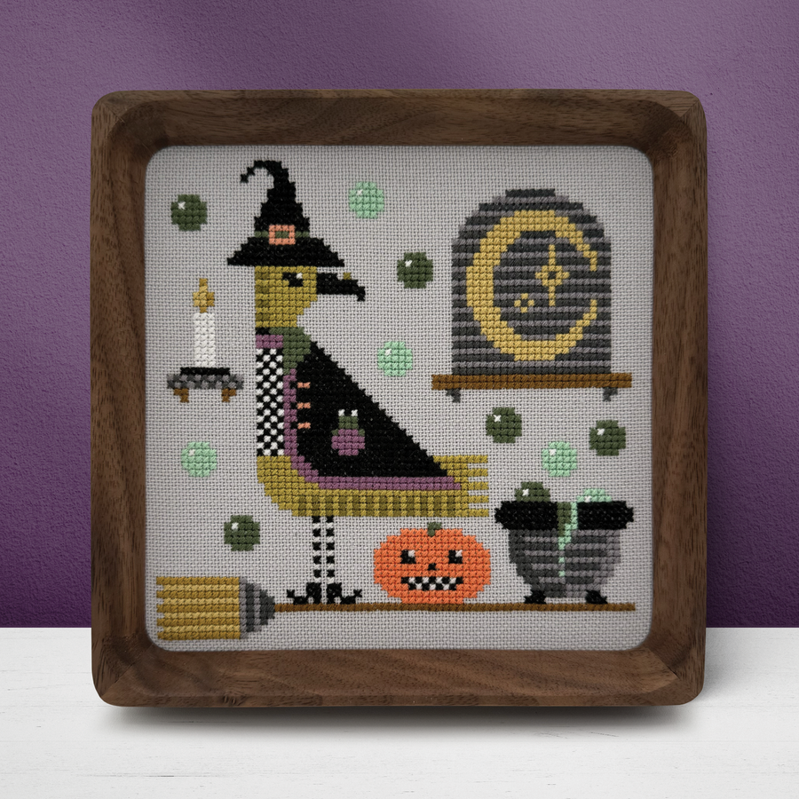 Halloween-themed cross-stitched design with a witch, pumpkin, and broomstick in a wooden frame against a purple background.