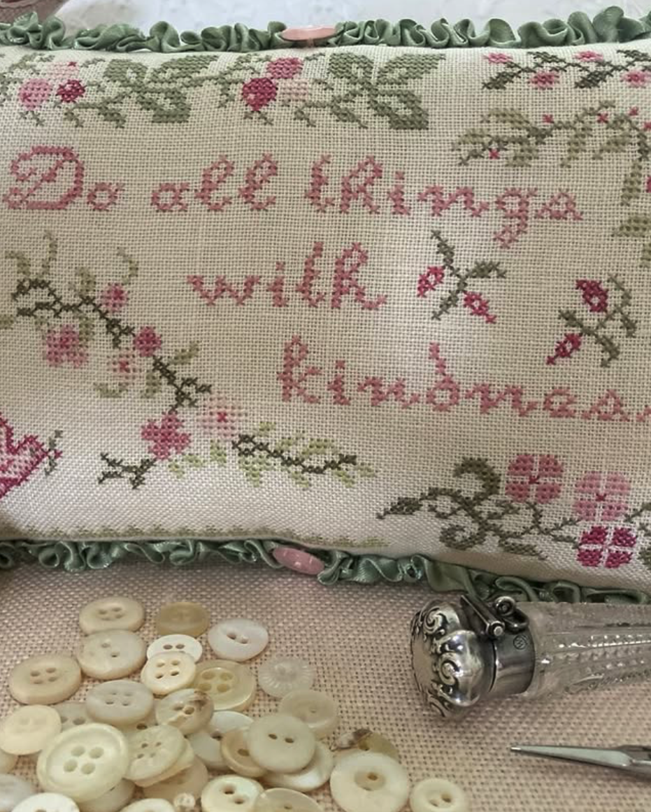 Cross-stitched pillow with floral patterns and text, surrounded by buttons and a thimble on a fabric background.