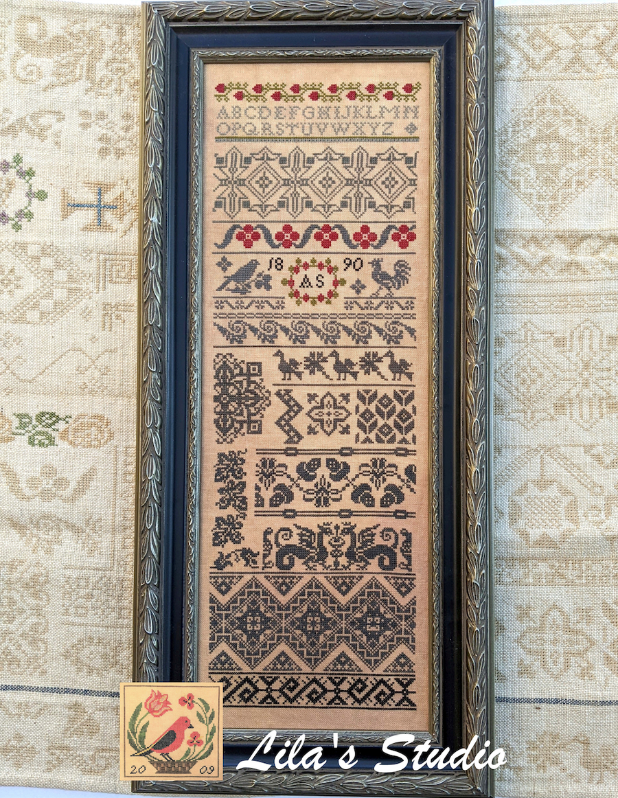 Framed cross-stitch sampler with decorative border on a textured fabric background, featuring 'Lila's Studio' branding.