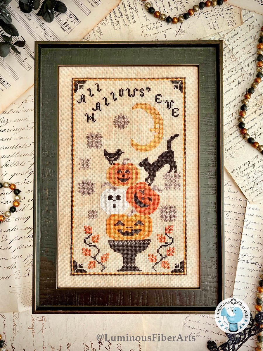 Framed Halloween-themed cross-stitch art with pumpkins, cats, and text on a textured background.