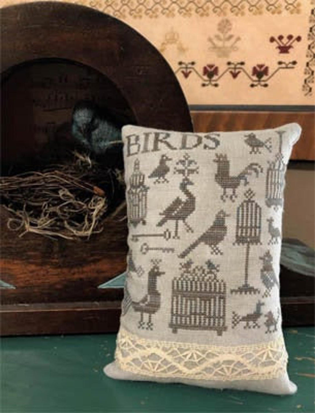 A cross-stitch pillow with bird and cage designs, the word 'BIRDS' across the top, displayed on a wooden surface.