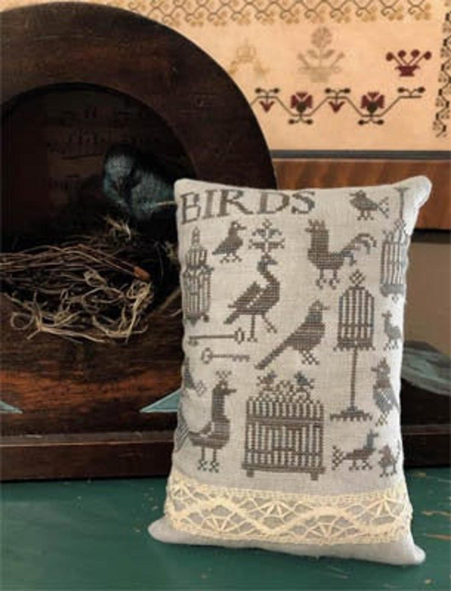 A cross-stitch pillow with bird and cage designs, the word 'BIRDS' across the top, displayed on a wooden surface.