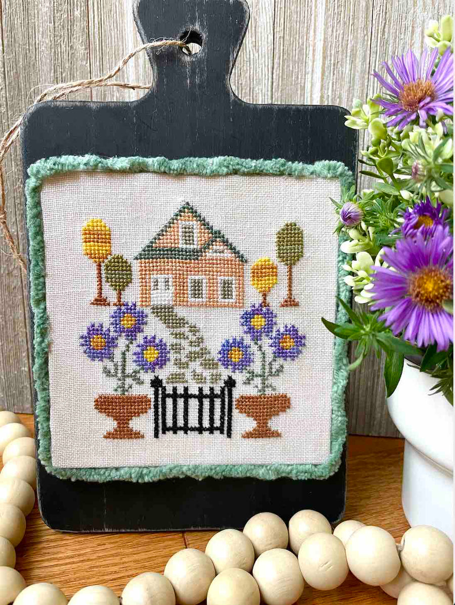 Cross-stitched house with flowers on a wooden stand with decorative elements.