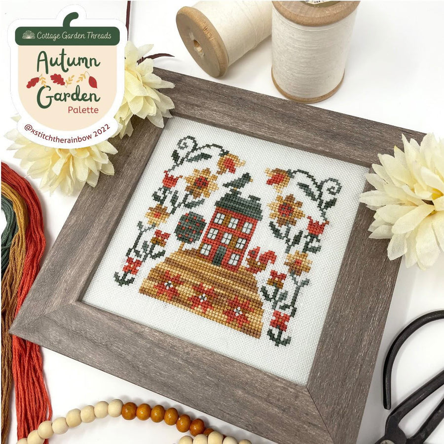 Framed cross-stitched piece with autumn theme on a white background with thread and flowers.