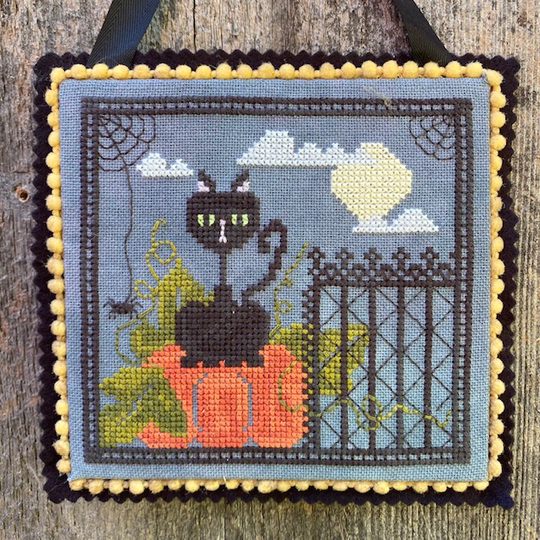 Cross-stitched picture of a black cat on pumpkins with a fence and moon in the background.