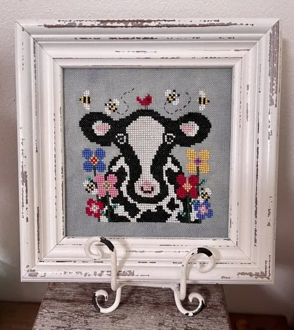 Framed cross-stitched cow with flowers and bees on a gray background