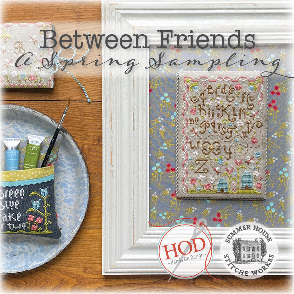 Cross-stitched sampler with 'Between Friends' text, surrounded by embroidery supplies on a wooden surface.