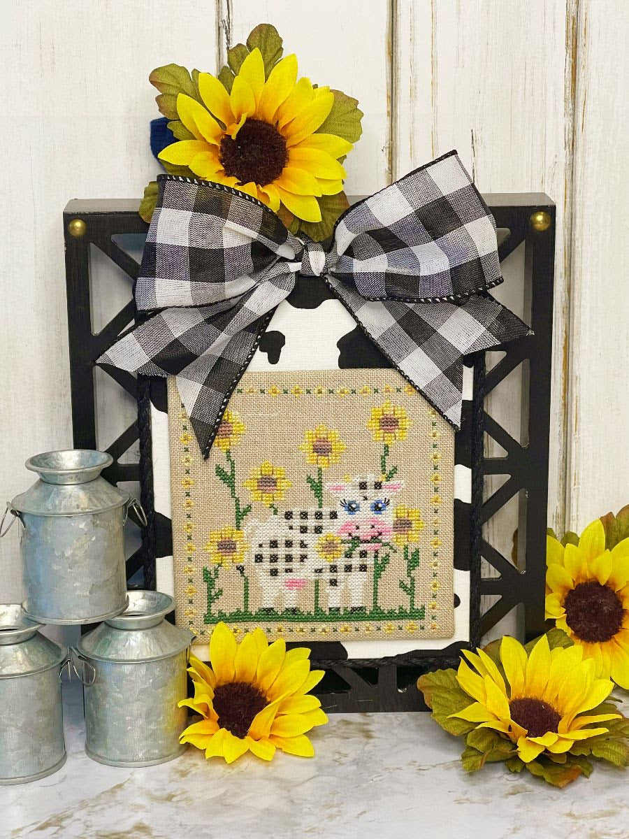 Decorative cross-stitched piece with sheep and sunflowers, framed with a checkered bow, surrounded by sunflowers and small metal buckets.