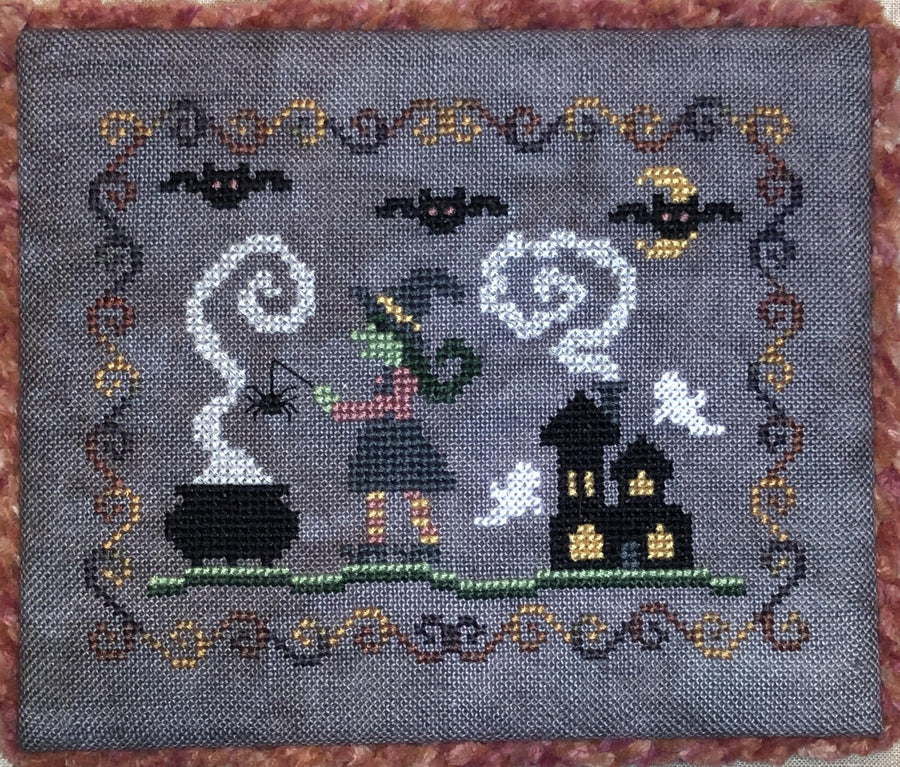A square cross-stitch sampler featuring a Halloween theme on a gray fabric background. The design includes a witch with  a smoking cauldron, a haunted house, bats, a crescent moon, and ghosts, framed by a decorative border.
