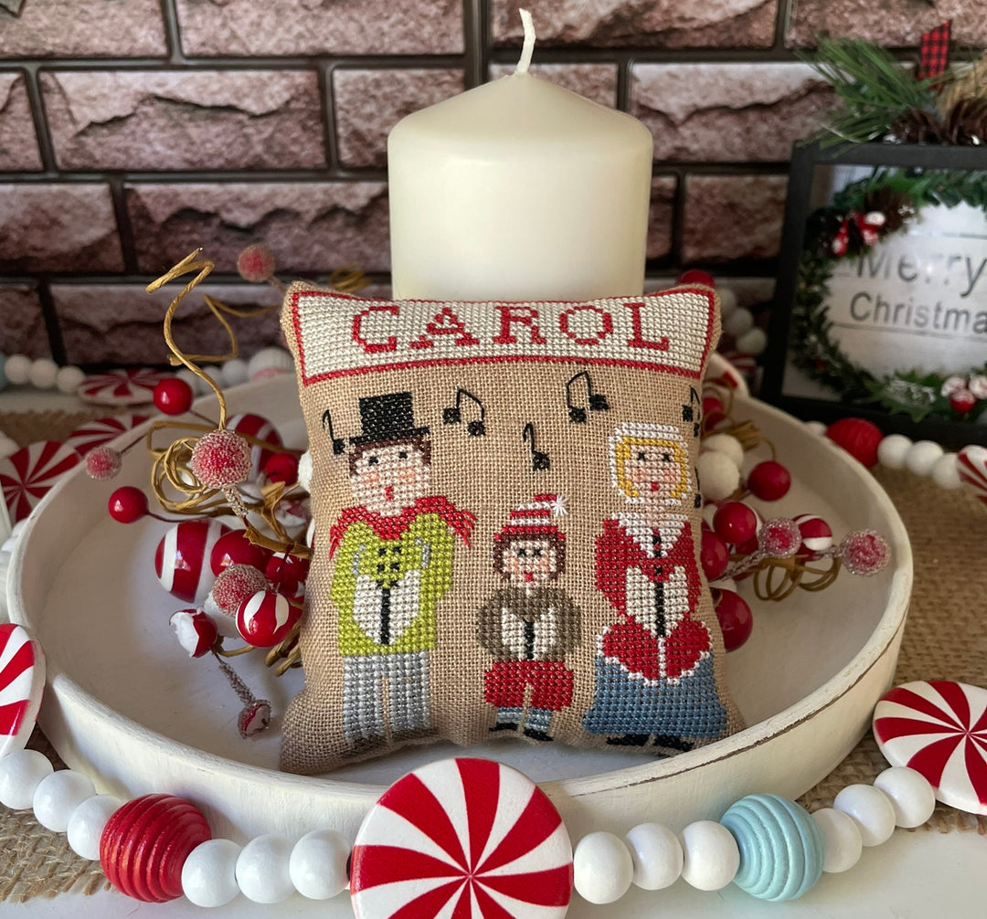 Decorative pillow with 'CAROL' text, surrounded by Christmas decorations on a stone wall background.