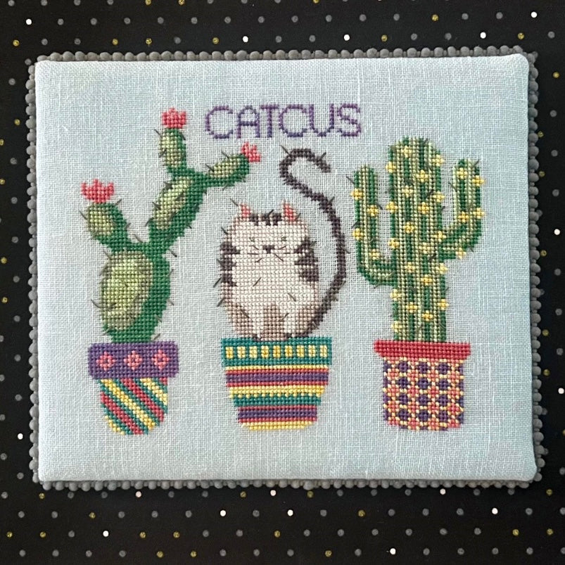 Cross-stitched design with a cat and two cacti on a light blue background