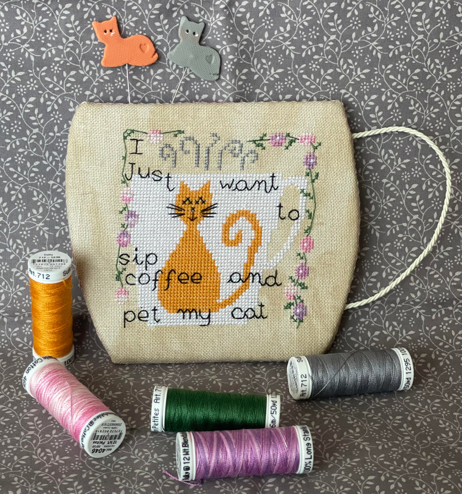 Cross-stitched pouch with a cat design and text, surrounded by spools of thread on a patterned background.