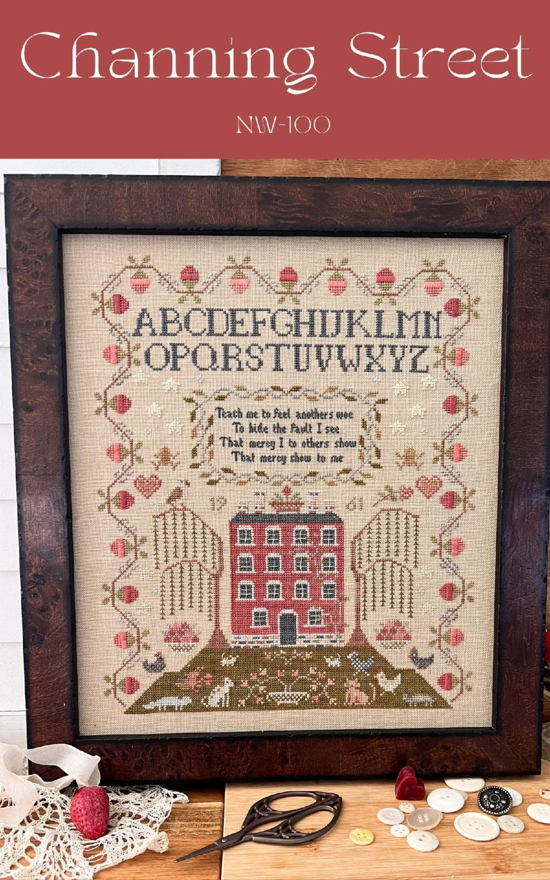 A framed needlework piece with the alphabet, a house, and decorative elements, set on a table with embroidery scissors and a hoop.