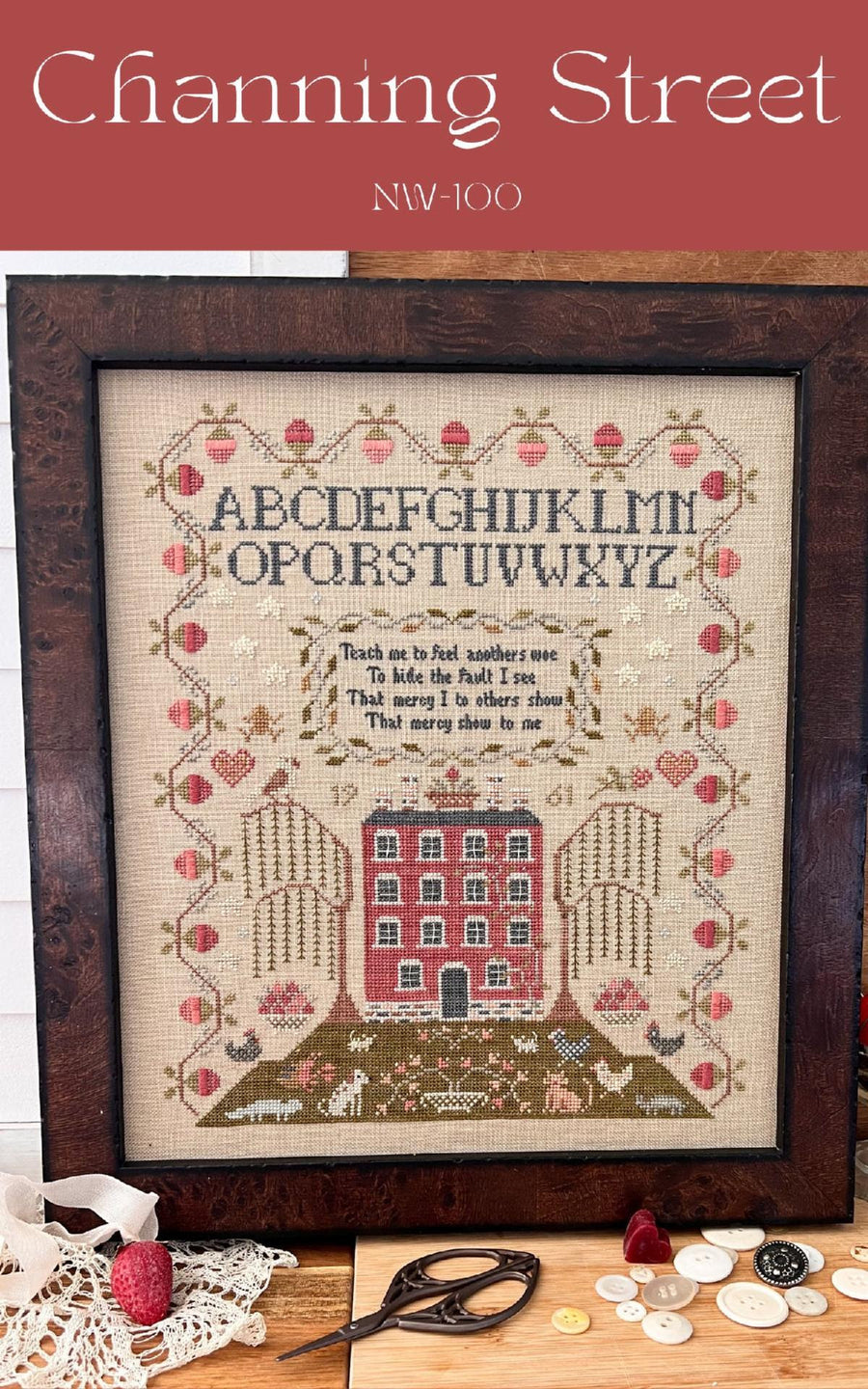 A framed needlework piece with the alphabet, a house, and decorative elements, set on a table with embroidery scissors and a hoop.