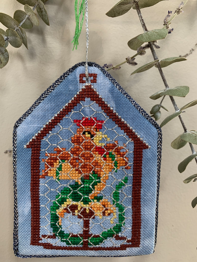 Decorative needlepoint ornament of a house with a rooster on a branch