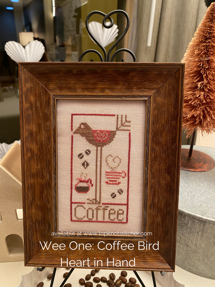 Framed cross-stitched artwork with a coffee-themed design on a table.