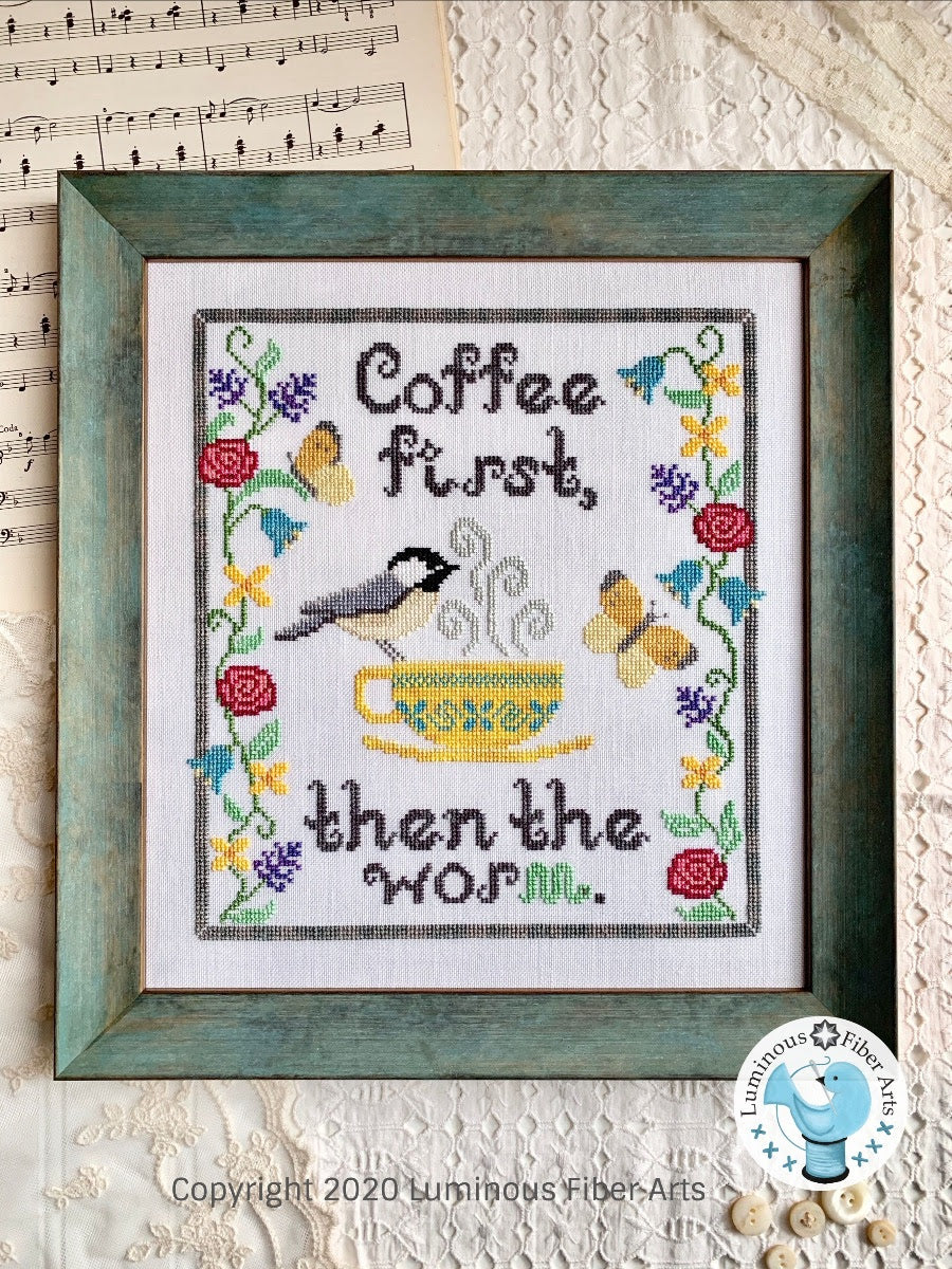 A needlecraft canvas with the text 'COFFEE FIRST, THEN THE WORLD' and an illustration of a bird (chickadee) perched on a cup of coffee, surrounded by floral patterns and the logo of Luminous Fiber Arts.