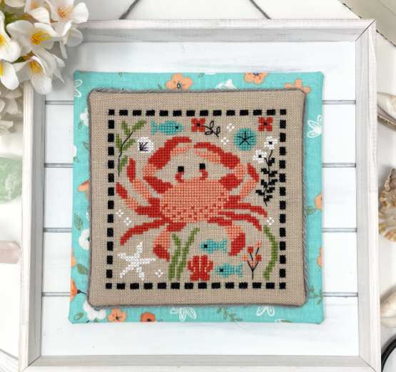 Framed cross-stitched crab design with floral elements on a white wooden surface.