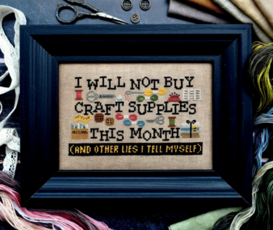 Cross-stitch kit depicting a humorous phrase about not buying craft supplies this month, surrounded by craft-related items like thread and scissors, framed in a black frame.