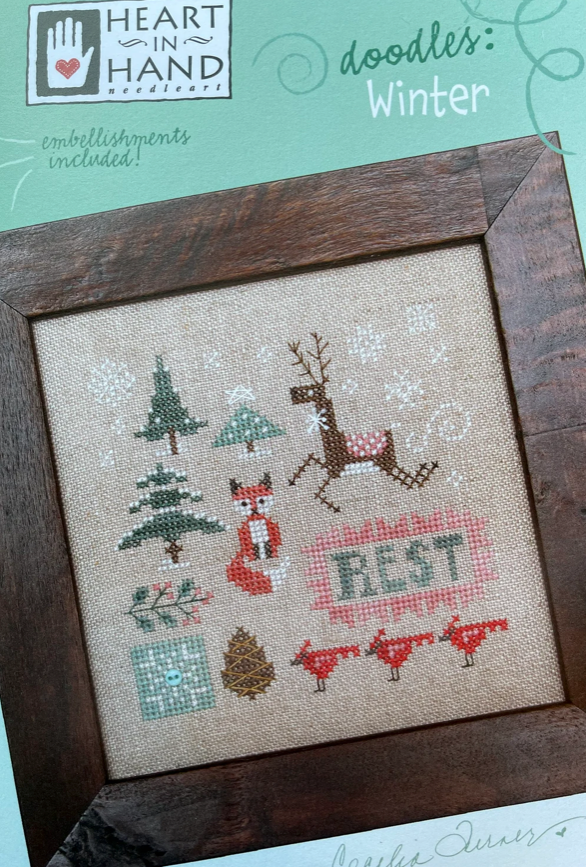 Framed cross-stitched design with winter-themed elements on a textured fabric background.