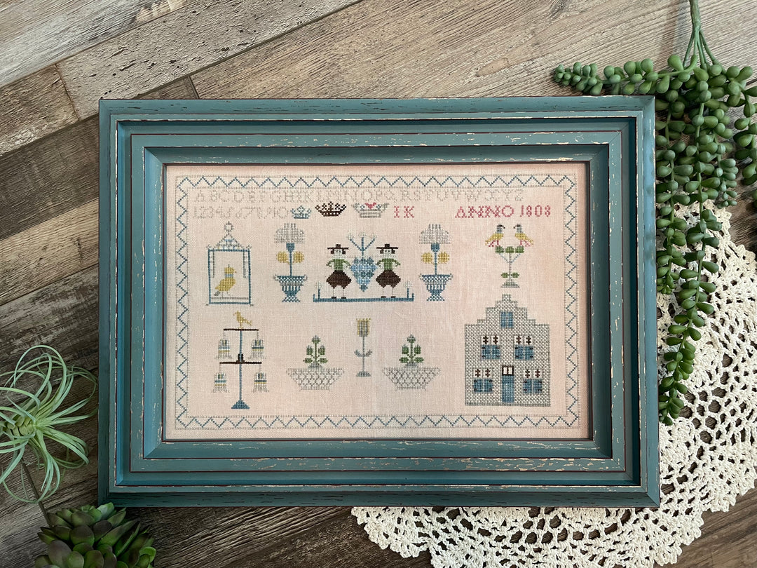 Framed cross-stitched sampler on a wooden surface with plants and lace