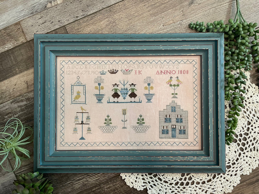 Framed cross-stitched sampler on a wooden surface with plants and lace