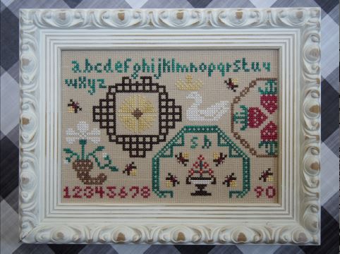 Framed cross-stitched sampler with alphabet, numbers, and decorative elements on a checkered background.