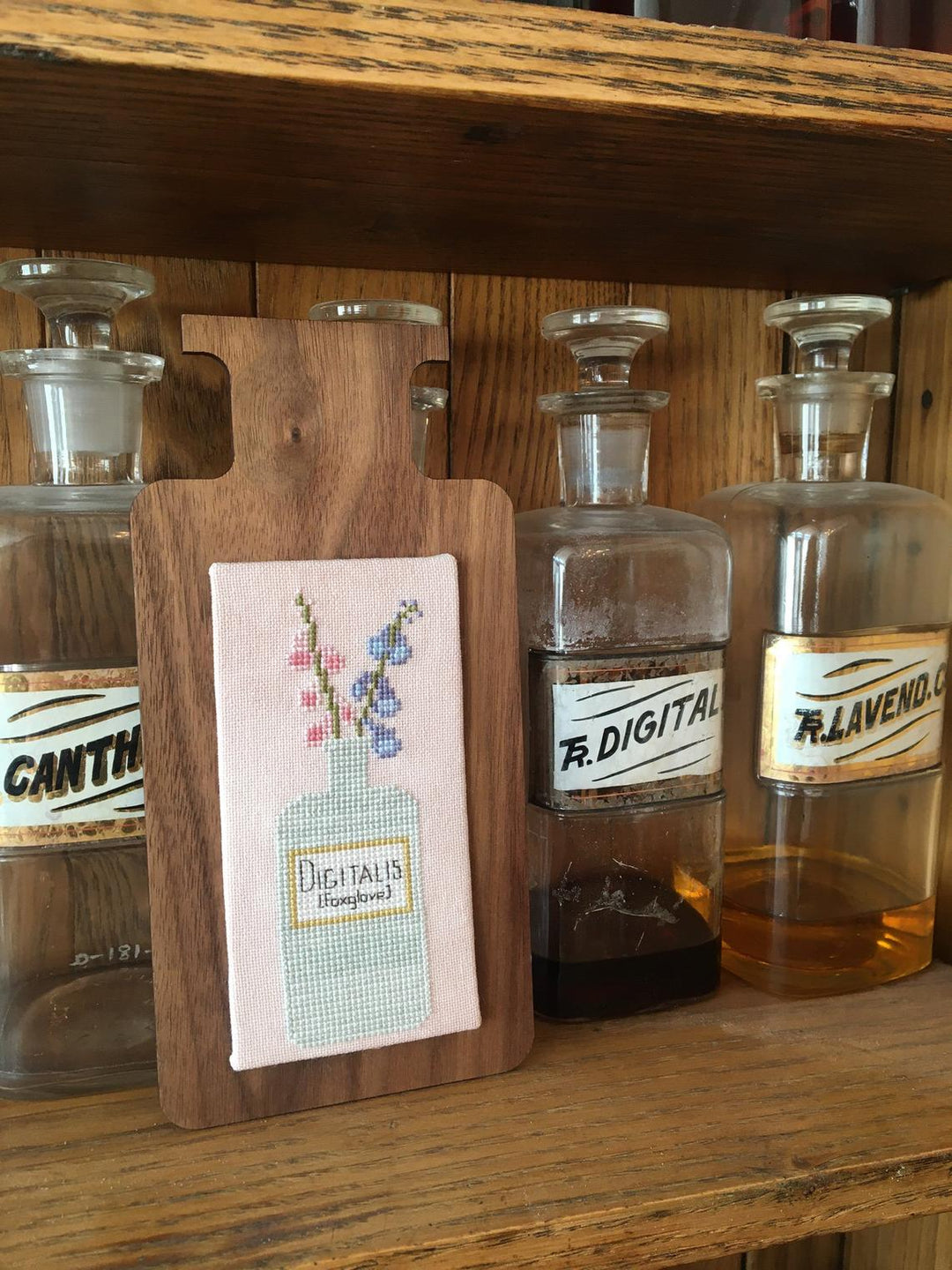 Wooden cutting board with cross-stitched design of a bottle with flowers, placed on a wooden shelf with glass bottles.