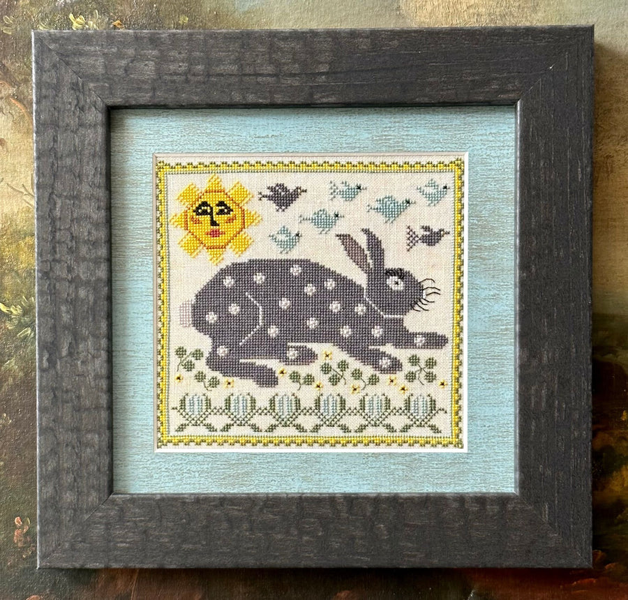 Framed cross-stitched artwork of a rabbit with a sun and birds on a textured background
