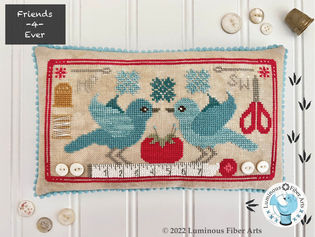Cross-stitched pillow with two blue birds, a red tomato, and a pair of scissors on a white background.