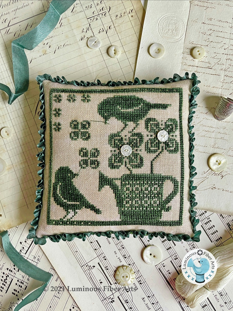 Decorative pillow with cross-stitch design of birds and a teacup on a textured surface.