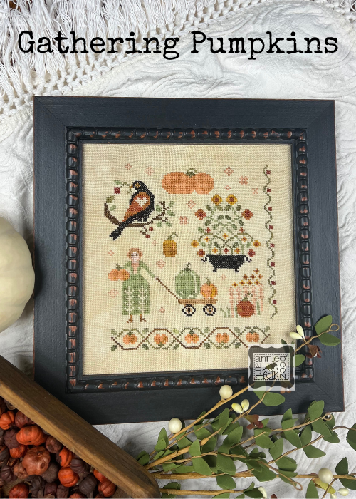 framed cross stitch featuing a blackbird flwoers woman and wagon with multiple pumpkins 