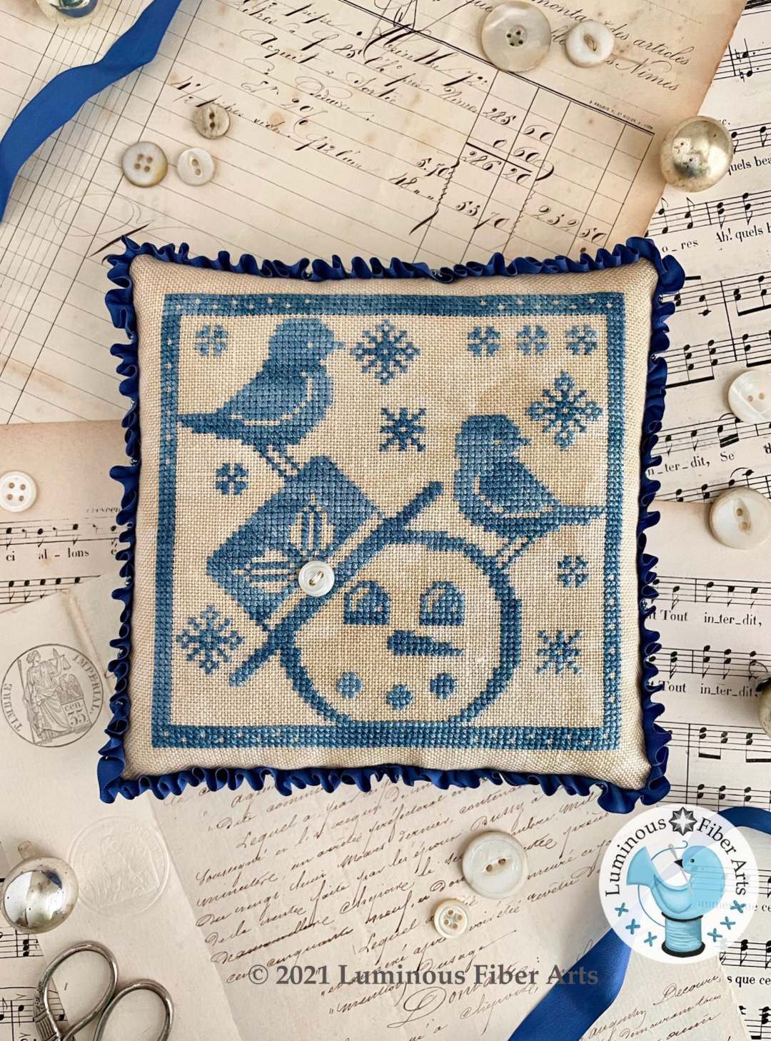 Cross-stitched pillow with bicycle and bird design on a vintage paper background, featuring Luminescent Fiber Arts logo.