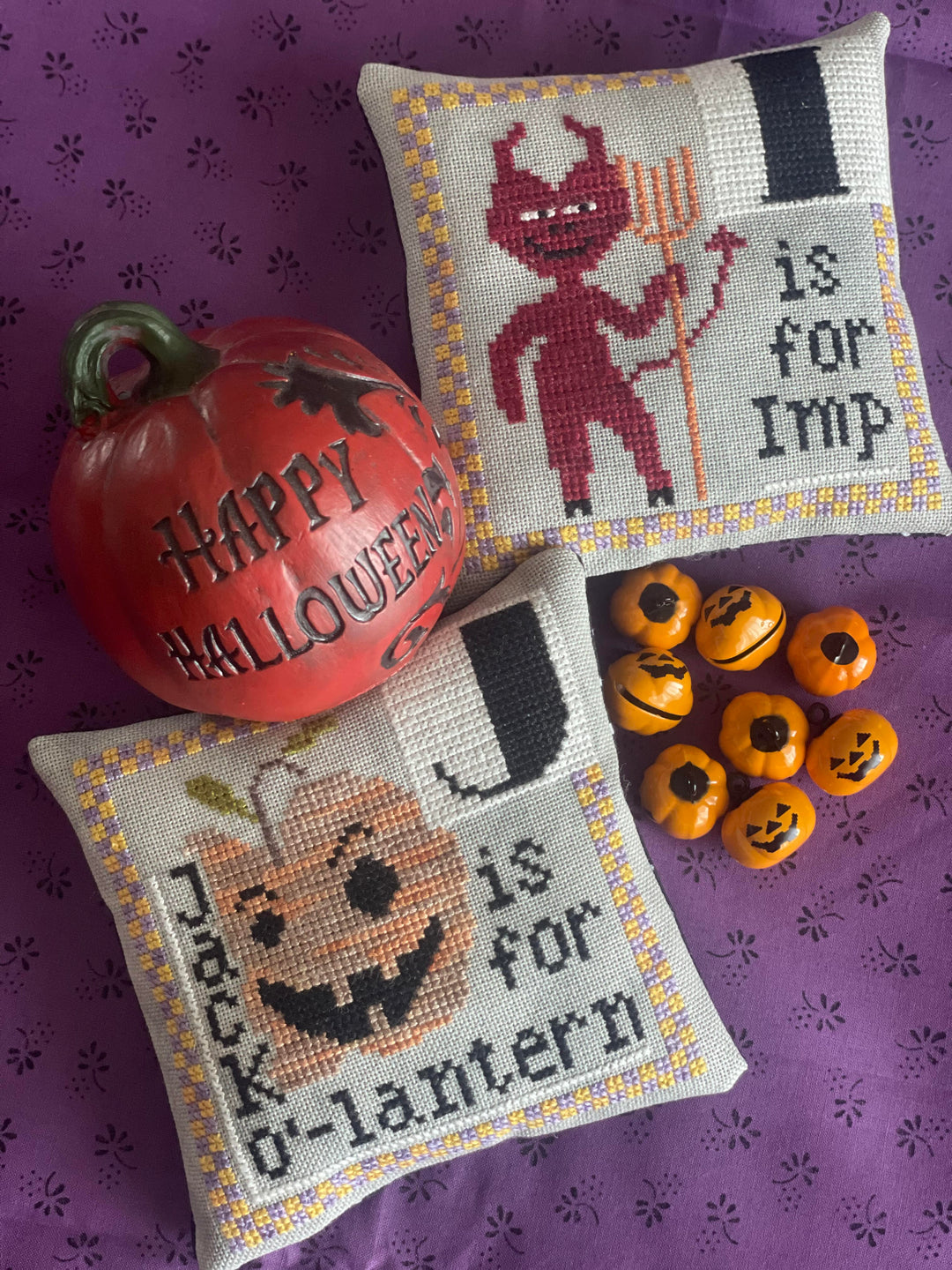 Two Halloween-themed cross-stitched pillows with decorative pumpkins and small jack-o'-lanterns on a purple background.