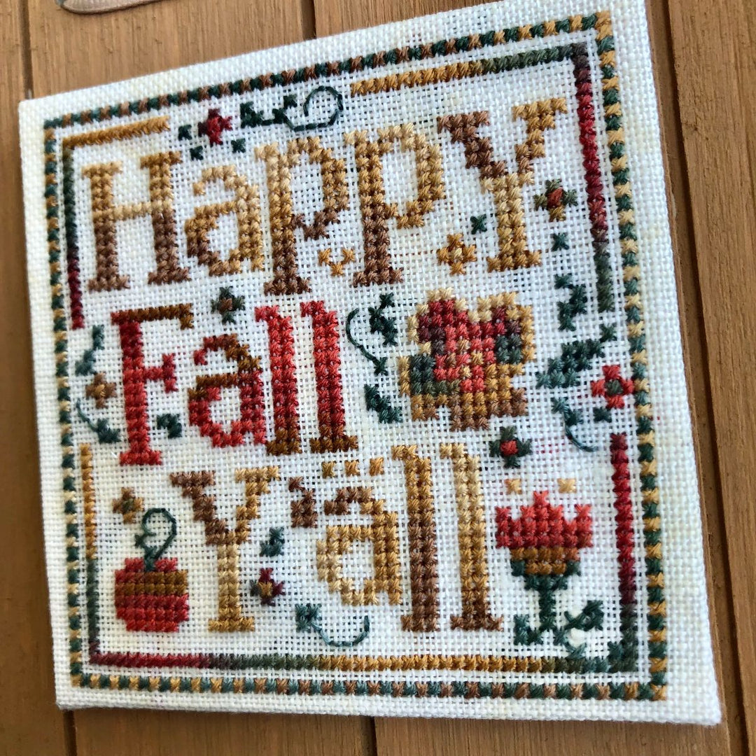 Cross-stitched sampler with 'Happy Fall Y'all' text and autumnal elements on a wooden surface.