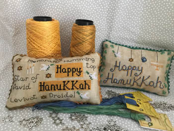 Two square pillows with 'Happy Hanukkah' text and decorative patterns, one with a yellow spool of thread, on a white background.