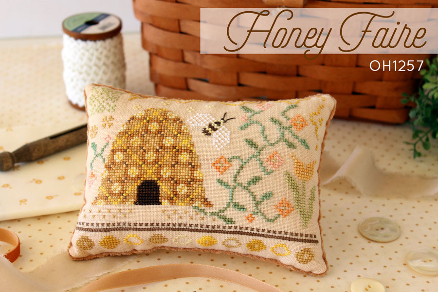 A cross-stitch depiction of a bee skep set among flowers, with a variety of stitches and colors used to create the design.