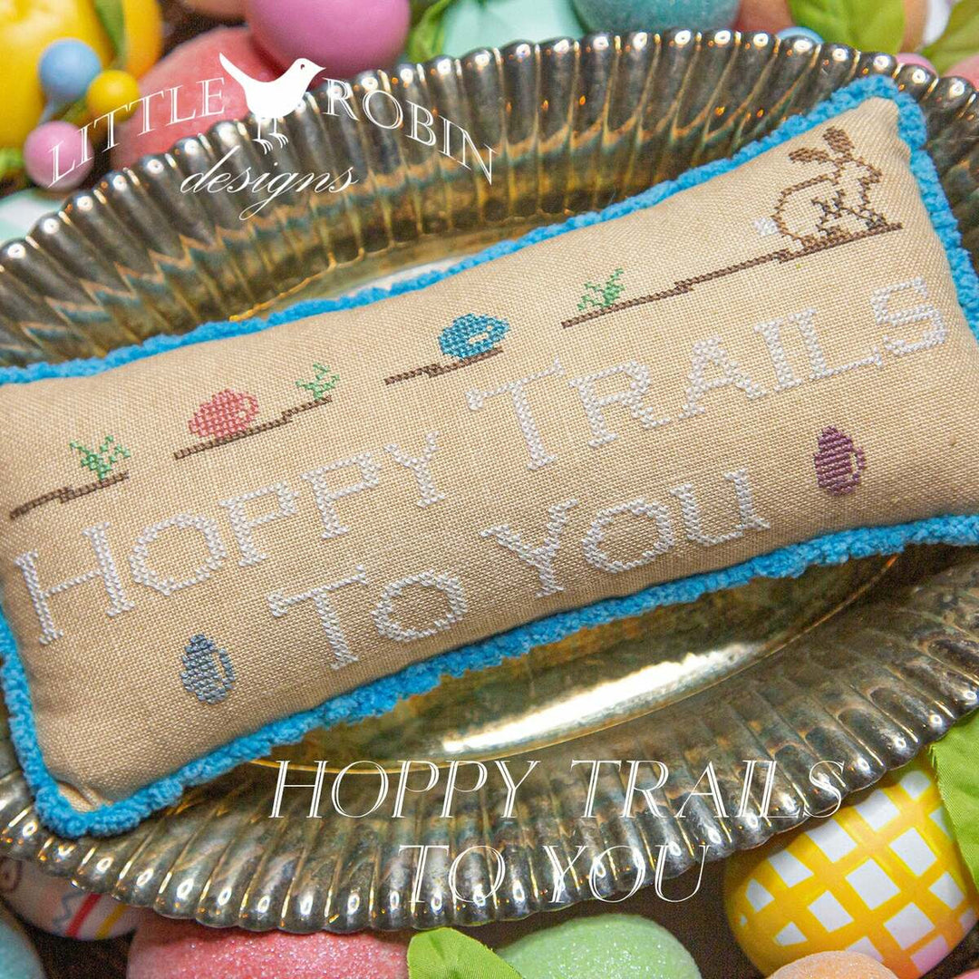 Cross-stitch needlework with the phrase 'Hoppy Trails To You' surrounded by stylized images of a bunny and Easter eggs, presented on a plate.