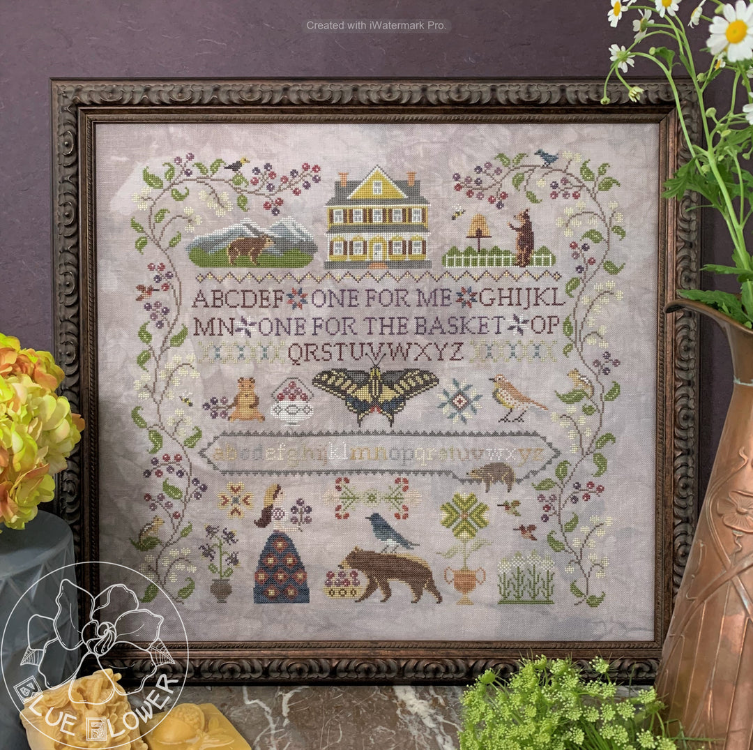 A cross-stitch pattern depicting a rural scene with a big house, trees, and a blue flower, surrounded by decorative elements.
