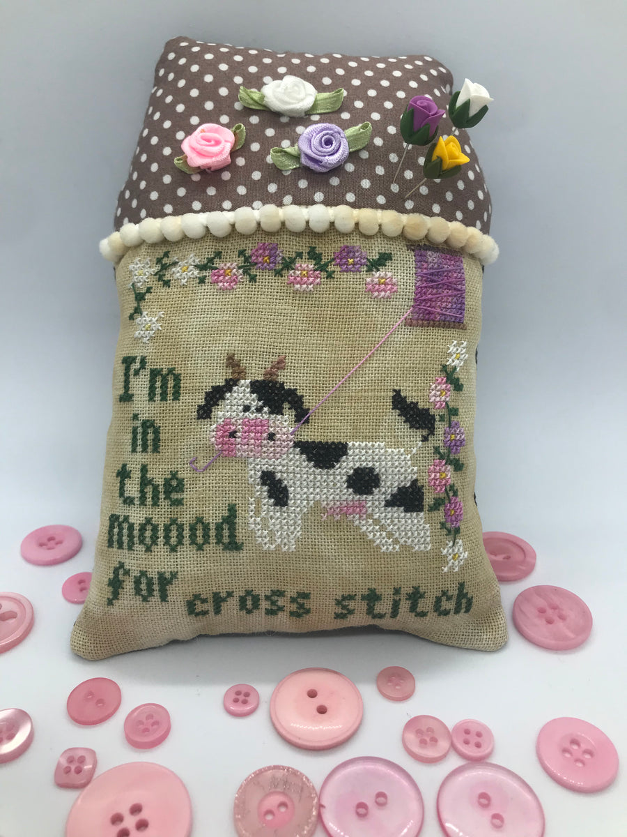 Decorative pillow with cross-stitched design of a cat holding a needle and thread, surrounded by pink buttons on a light gray background.