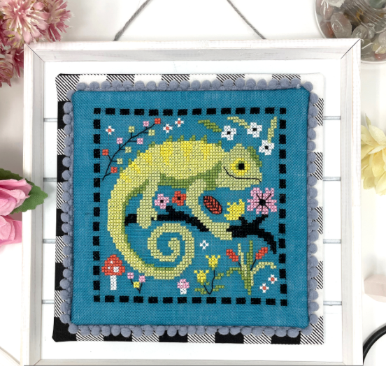Cross-stitched chameleon design on fabric with a white frame and floral decorations.