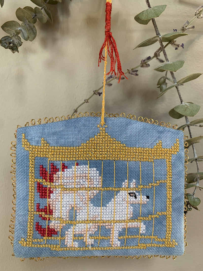 Decorative needlepoint hanging with animals in a cage design, hanging from a branch.