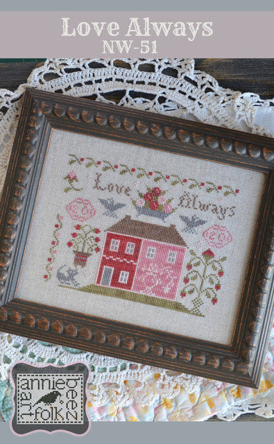 A framed cross stitch needlework depicting a red house with the phrase 'Love Always' displayed at the top.