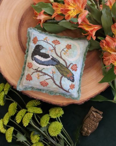 Decorative needlepoint pillow with a bird design on a wooden surface surrounded by flowers.