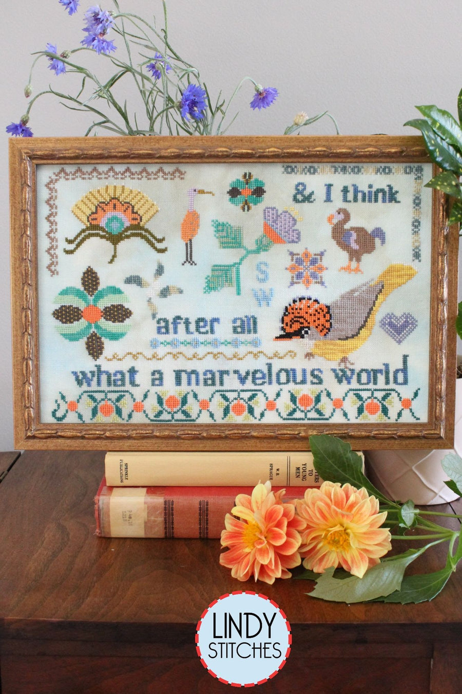Framed cross-stitched artwork with floral and nature-themed design, books, and flowers on a wooden surface.