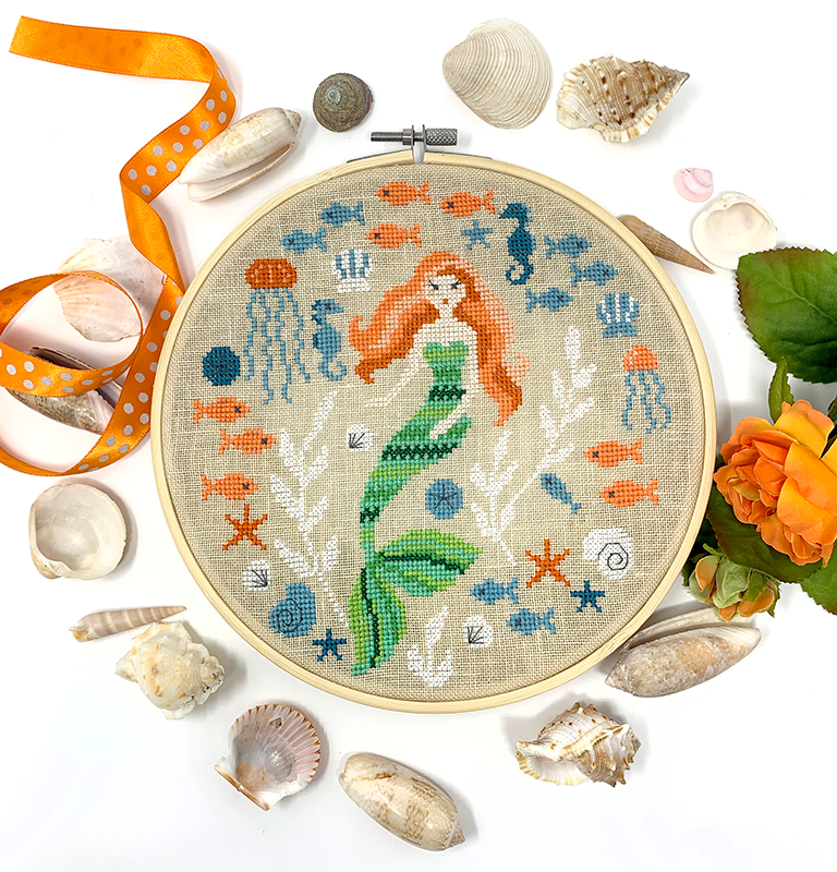 Embroidery hoop with mermaid design surrounded by shells and flowers on a white background