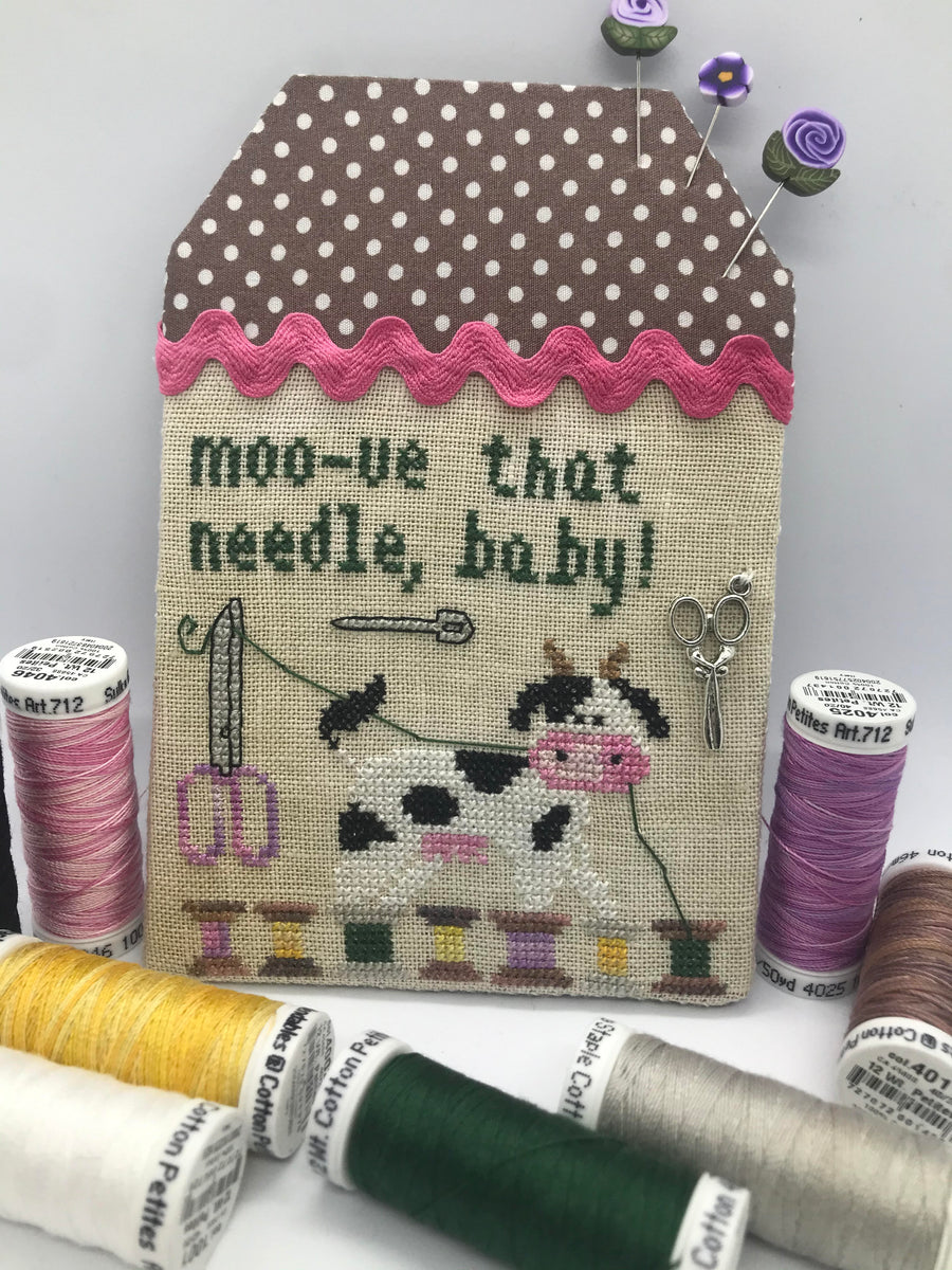Decorative needle case with cow illustration and text, surrounded by spools of thread.